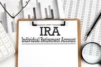 A stock image of a paper on a clipboard reading &quot;IRA&quot; and &quot;Individual Retirement Account&quot; on a table with other papers.