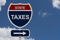 The words &quot;state taxes&quot; appear on a red, white, and blue, shield-shaped sign that imitates an interstate sign, against a blue sky and wispy white clouds.