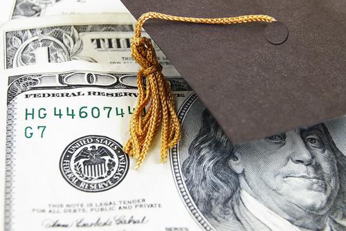 A corner of a miniature mortarboard lays on an array of $100 bills